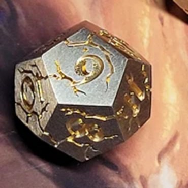 D12 — Spiral Galaxy die, full scale: a brushed silver metal twelve-sided die with a gold spiral galaxy engraved across its faces, cropped from the set photo