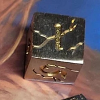 D6 — Constellation Cube die, full scale: a brushed silver metal six-sided cube with gold constellation lines and stars engraved across its faces, cropped from the set photo